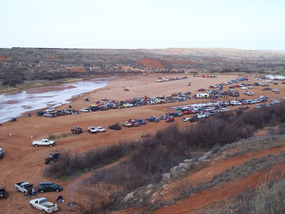 Many cars and trucks parked on a dirt area near a river, with a train visible on a distant ridge
