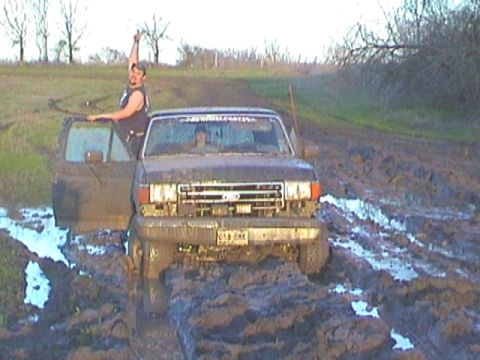 1989 Ford Bronco - A 1989 Ford Bronco is stuck in deep mud, with a person in the driver's seat and another leaning out the door giving a thumbs up.
