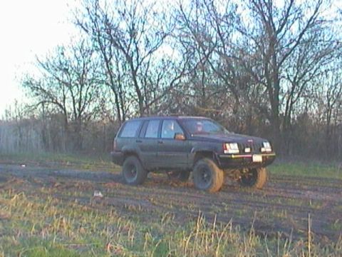 1989 Ford Bronco - A dark Jeep Grand Cherokee is shown from a 3/4 front angle on a muddy road with trees in the background.