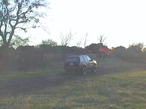 1989 Ford Bronco - A dark SUV is seen driving through a grassy field with trees in the background.