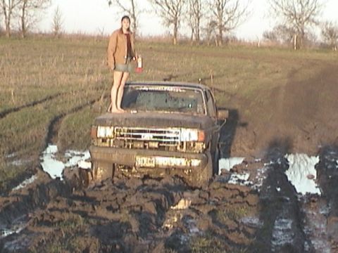 1989 Ford Bronco - A woman stands on the hood of a mud-covered 1987 Ford F-Series truck stuck in a muddy field.