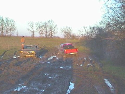 1989 Ford Bronco - Two trucks are stuck deep in the mud, with a person standing on the hood of the brown truck.
