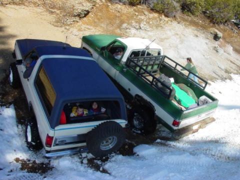1986 Ford Bronco - Elevated rear 3/4 view of a white and blue lifted Ford Bronco on mud-terrain tires and polished wheels, with kids waving through the open r…
