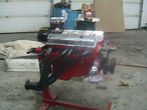 1981 Chevrolet C/K Pickup - A red V8 engine with chrome valve covers and black headers is displayed on a stand in a garage setting.