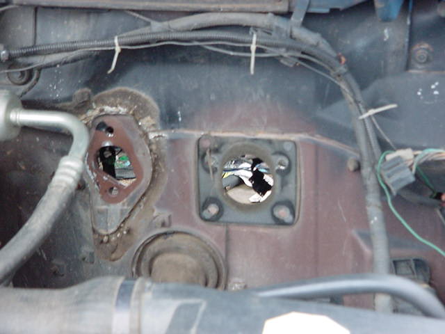 Firewall with two large holes, one with a brown gasket, near AC line and wiring harness