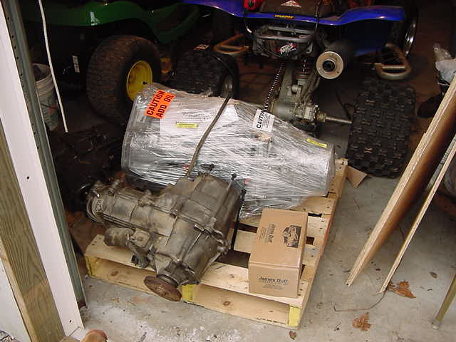 Transfer case and transmission parts on wooden pallets in a garage. A blue ATV and green lawn tractor are visible in the background
