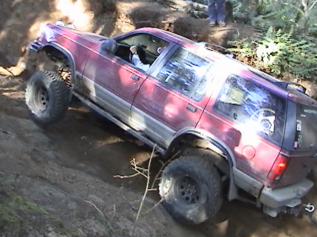 Red SUV with large tires climbing a dirt incline. The vehicle has a two-tone lower body panel and roof rack
