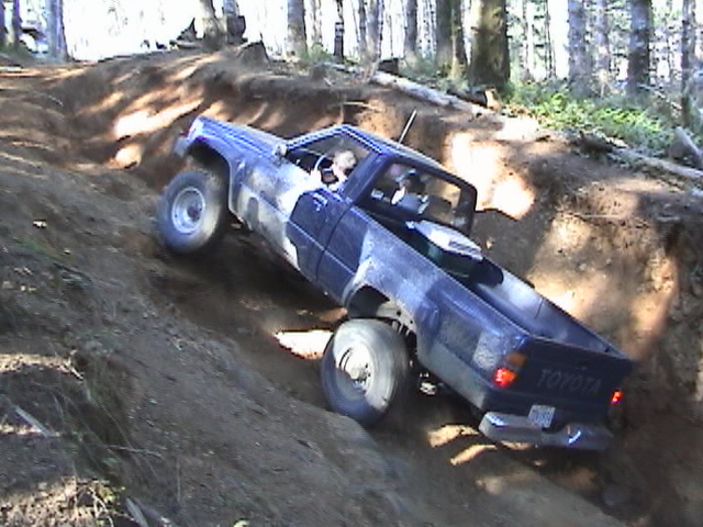 Blue chopped-top Toyota pickup climbing out of a dirt trench on a wooded off-road trail