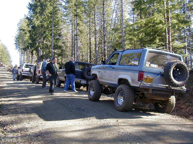 Several off-road SUVs parked on a dirt road in a forest, with people standing around them