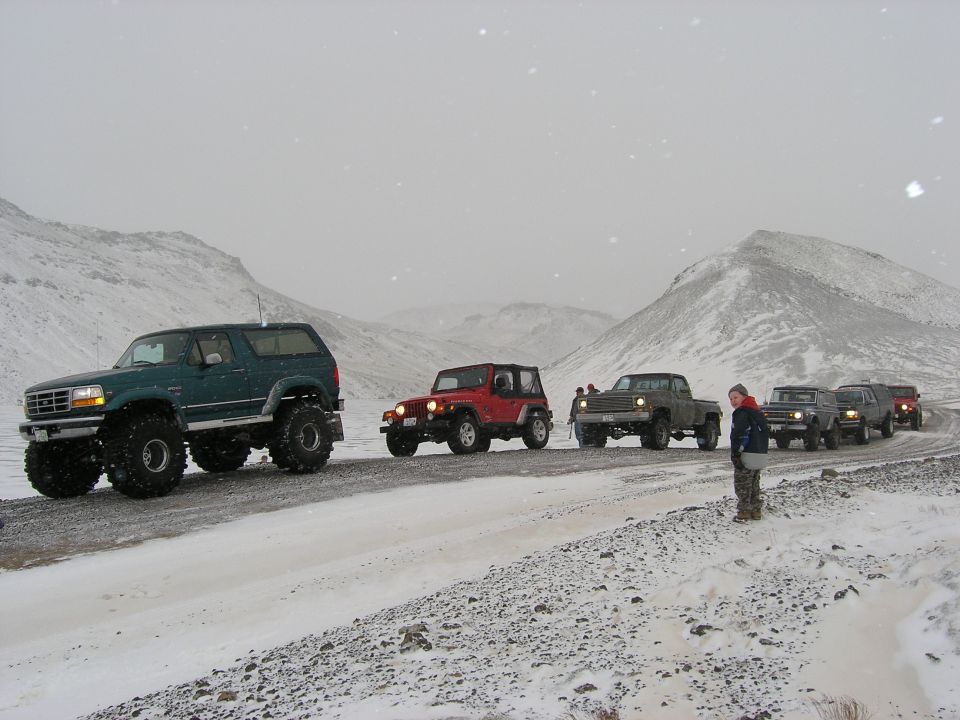 Several off-road vehicles driving on a snowy road with mountains in the background. One person stands by the side of the road