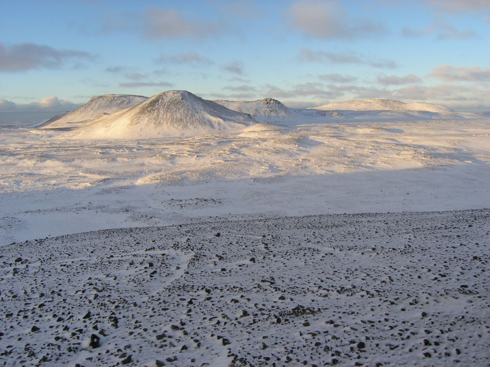 Snow-covered terrain with rolling hills and mountains under a blue sky with clouds