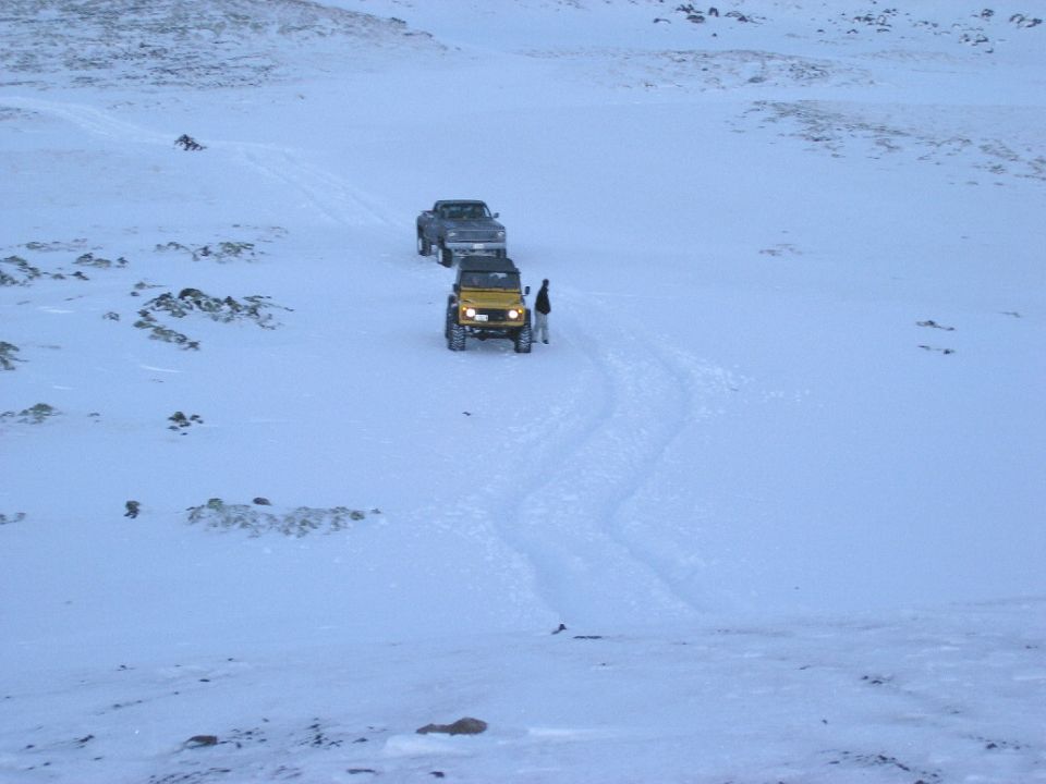 Yellow Jeep and gray pickup truck driving on a snowy trail with two people standing near the Jeep