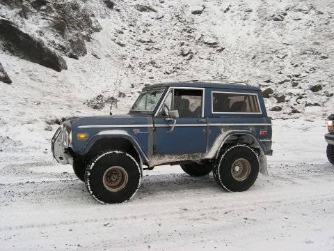 2001 Jeep Wrangler - A blue Ford Bronco is parked on a snowy surface with a rocky, snow-covered hill in the background.