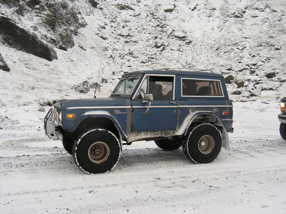 Blue Ford Bronco with big tires on snowy ground, another vehicle partially visible to the right
