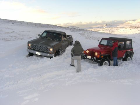2001 Jeep Wrangler - Two vehicles, a red Jeep and a dark pickup, are stuck in a snowy landscape with two people observing.