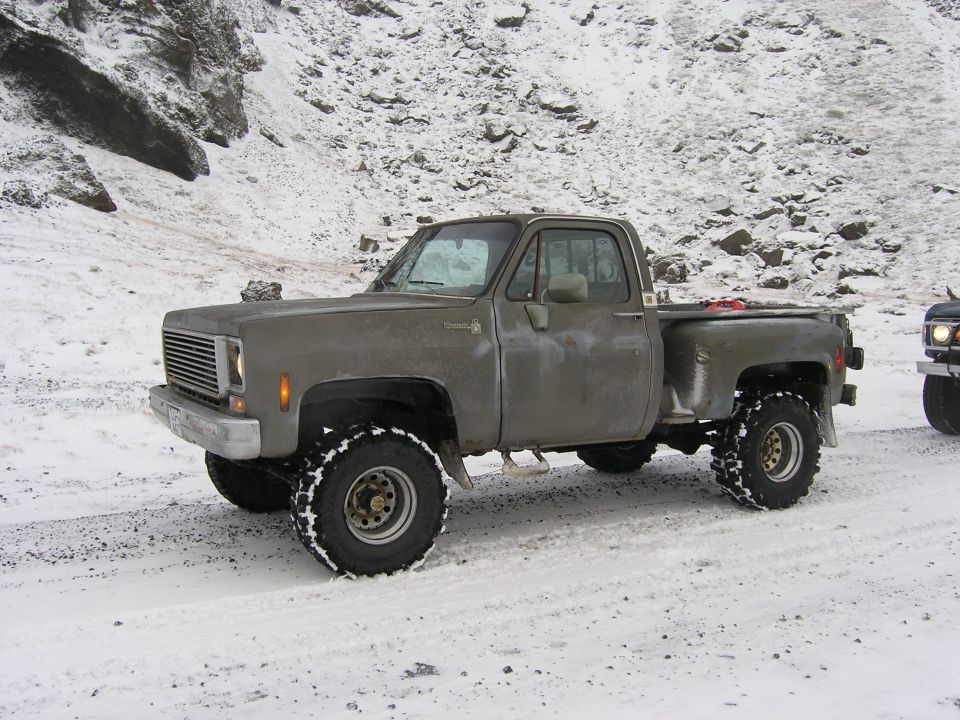 Old pickup truck with large tires on a snowy road, another vehicle visible to the right