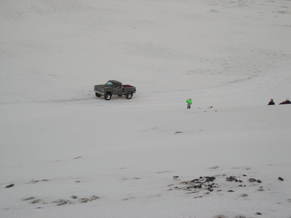 Gray pickup truck driving on a snowy field with a person in a green jacket nearby