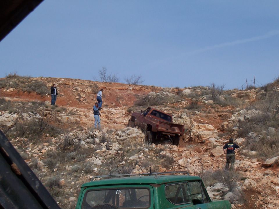 Red pickup truck struggling on rocky terrain with people watching