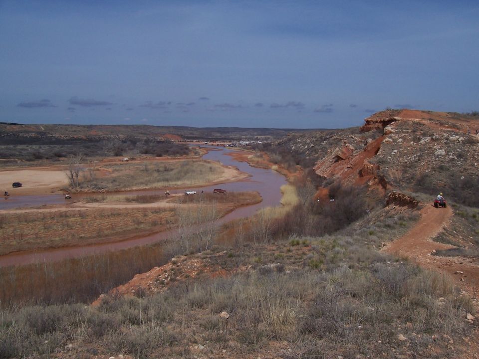 ATVs and cars driving in a muddy riverbed and on dirt trails in a dry, rocky canyon