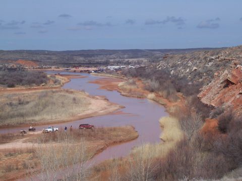 2004 Aston Martin Virage - A wide shot captures a muddy river winding through a desolate landscape with several vehicles parked along its banks.