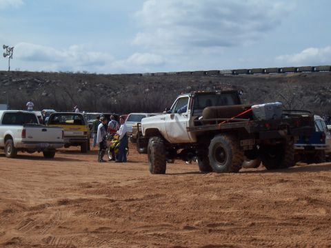 2004 Aston Martin Virage - A white flatbed truck with oversized tires is parked on a dirt lot alongside other vehicles and people.