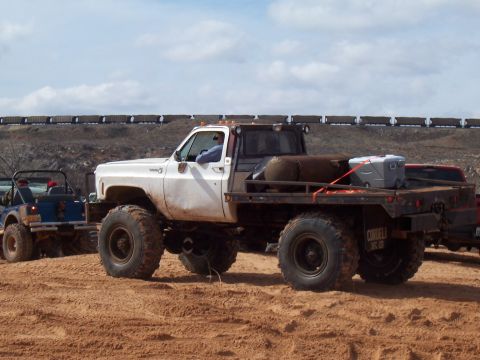 2004 Aston Martin Virage - A heavily modified flatbed truck with large tires is seen in a sandy area, with a train visible on a ridge in the distance.