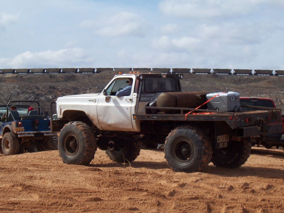 White flatbed truck with big tires, towing a blue jeep. Train in background