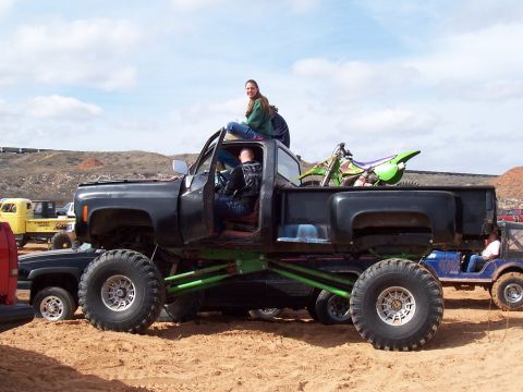 2004 Aston Martin Virage - A modified black truck with large tires and a dirt bike in the bed sits in sandy terrain with other vehicles nearby.