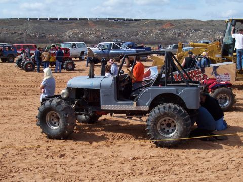 2004 Aston Martin Virage - A heavily modified jeep with oversized tires and a roll cage sits on sandy ground, with other vehicles and spectators in the background.