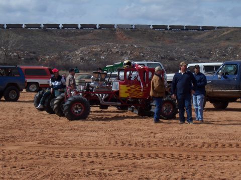 2004 Aston Martin Virage - Custom monster truck with flame decals and exposed engine sits in a sandy area with other vehicles and a train in the distance.