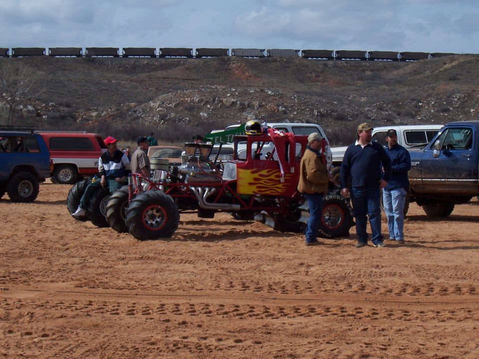 Custom red monster truck with yellow and red flames, large tires, and exposed engine. Several people stand around it. Other vehicles and a train are in the background