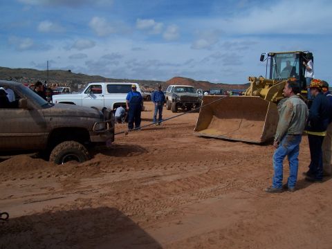 2004 Aston Martin Virage - A muddy pickup truck is stuck in the sand and being pulled by a large yellow front loader, with several people observing.