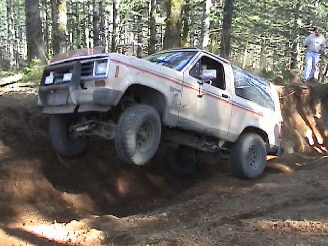 White Ford Bronco II climbing a dirt hill with trees in background. One person stands on a ledge
