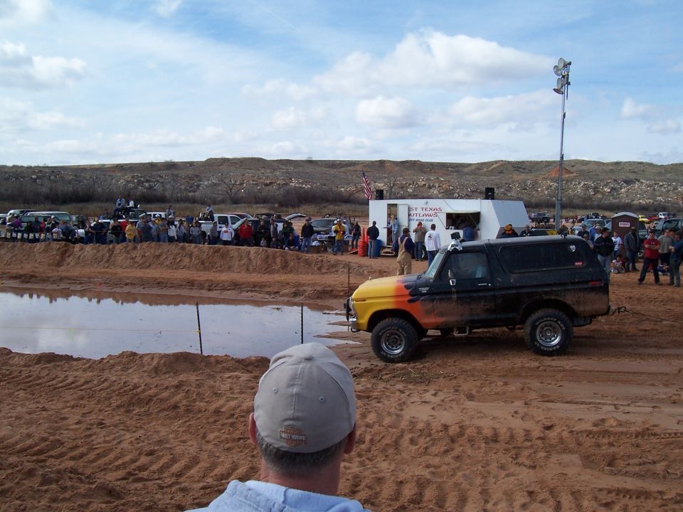 Ford Bronco with orange and yellow flame paint, parked at a mud bog event with spectators and other vehicles
