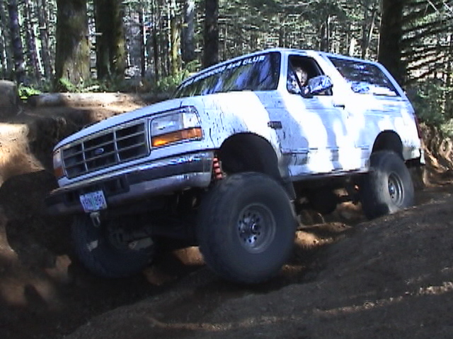 White Ford Bronco climbing a dirt hill in a forest, with a "BRONCO 4X4 CLUB" sticker on the windshield