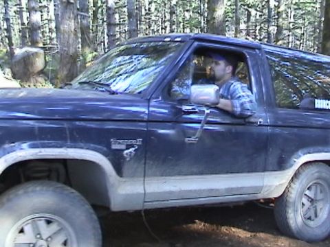 1994 Ford Explorer - A man drives a dark blue Ford Bronco II off-road through a wooded area.