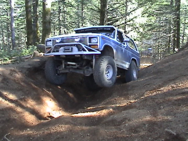 Blue Ford Bronco off-roading on a dirt trail, going over an obstacle. Another vehicle is visible behind it