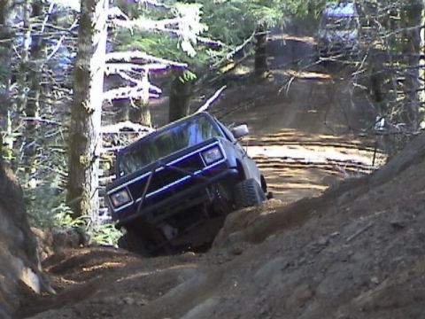 1994 Ford Explorer - An off-road vehicle navigates a challenging dirt trail in a wooded area, with another vehicle visible in the distance.