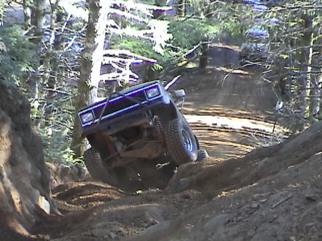 Off-road vehicle with lifted suspension climbing a steep dirt trail in a wooded area. Front grille and headlights are visible