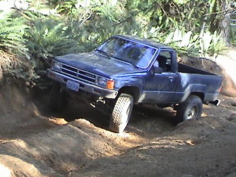 1994 Ford Explorer - A blue pickup truck is shown maneuvering on a challenging off-road trail surrounded by trees and dirt mounds.