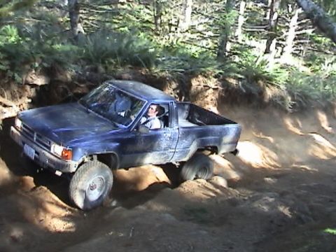 1994 Ford Explorer - A blue pickup truck is shown off-roading on a dusty trail with trees in the background.