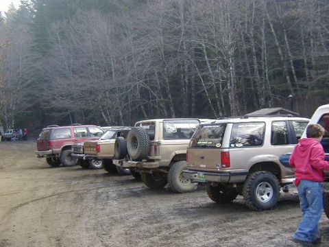 1994 Ford Explorer - Multiple SUVs are parked on a dirt road, with a forest in the background. One vehicle shows a spare tire on its rear.