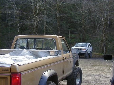 1994 Ford Explorer - Two off-road vehicles are parked in a dirt lot surrounded by trees. The white SUV has club text on its windshield.
