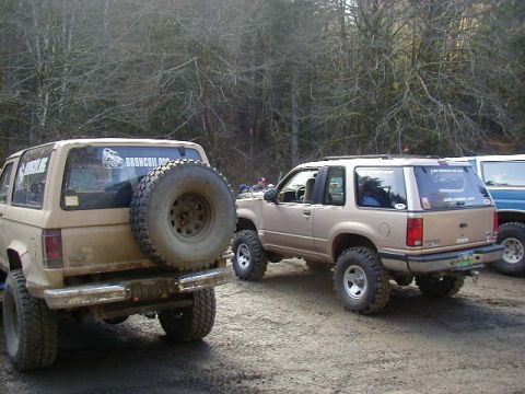 1994 Ford Explorer - Two off-road modified SUVs, a Ford Explorer and a Ford Bronco II, are parked on a dirt trail surrounded by trees.