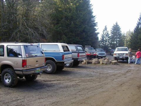 1994 Ford Explorer - Several SUVs and trucks are parked on a dirt lot near a forest, with people gathered around one of the vehicles.