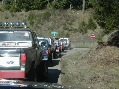 1994 Ford Explorer - A convoy of trucks and SUVs navigate a dirt road, pausing at a stop sign amidst a wooded hillside.