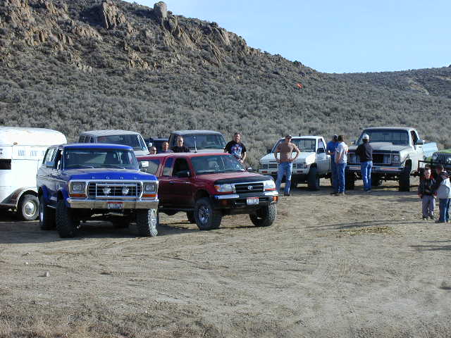 Group of lifted 4x4 trucks including a blue Bronco and red Toyota parked at desert trailhead