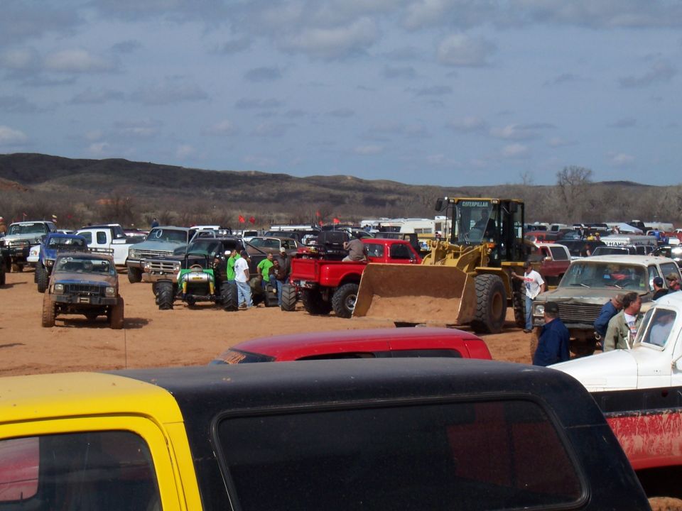 Many trucks, ATVs, and a front-end loader parked on a dirt lot under a cloudy sky