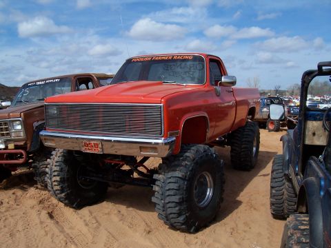 2004 Aston Martin Virage - A red lifted pickup truck with oversized tires and a racing banner sits in a muddy lot with other vehicles.