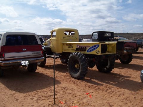 2004 Aston Martin Virage - A heavily modified yellow pickup truck with oversized tires sits on sandy ground next to a red SUV.